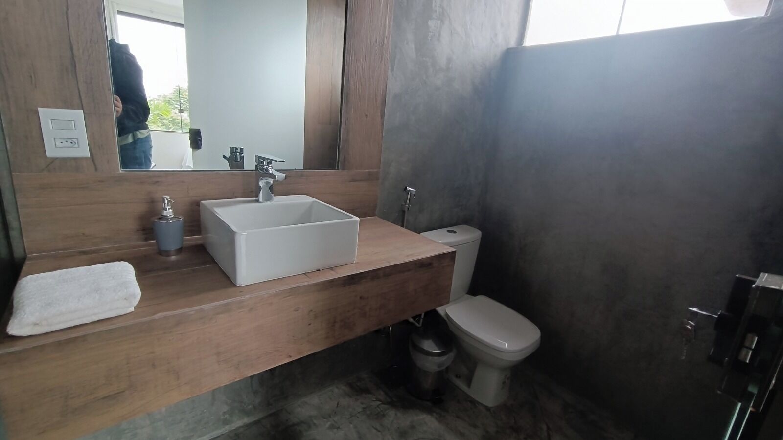A bathroom with a wooden vanity, a white sink, a mirror, a toilet, and a trash bin.