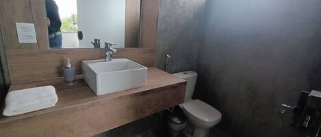 A bathroom with a wooden vanity, a white sink, a mirror, a toilet, and a trash bin.