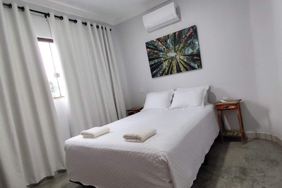 A neatly made bed with white linens and two folded towels, a bedside table with a lamp, and a wall-mounted air conditioner.