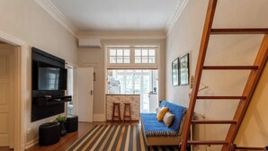 A living room with a TV, a sofa, a wooden ladder, and a kitchen visible through an open door.