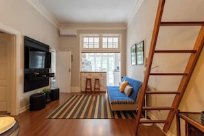 Ein Wohnzimmer mit Fernseher, einer Couch, einer Holzleiter und einer durchgehenden Tür erkennbaren Küche.