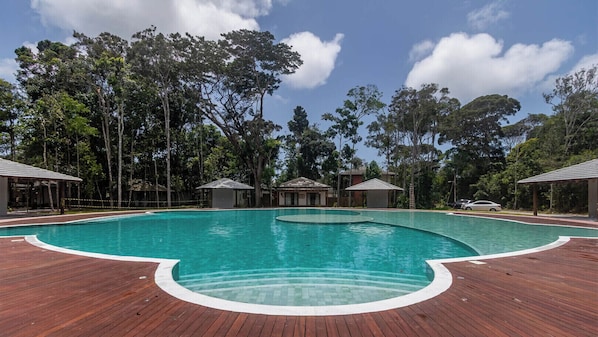 A large outdoor swimming pool with a wooden deck, surrounded by trees and covered structures.