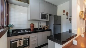 A modern kitchen with grey cabinets, a black countertop, a stainless steel refrigerator, and a microwave. There are decorative wall hangings and a small stool.