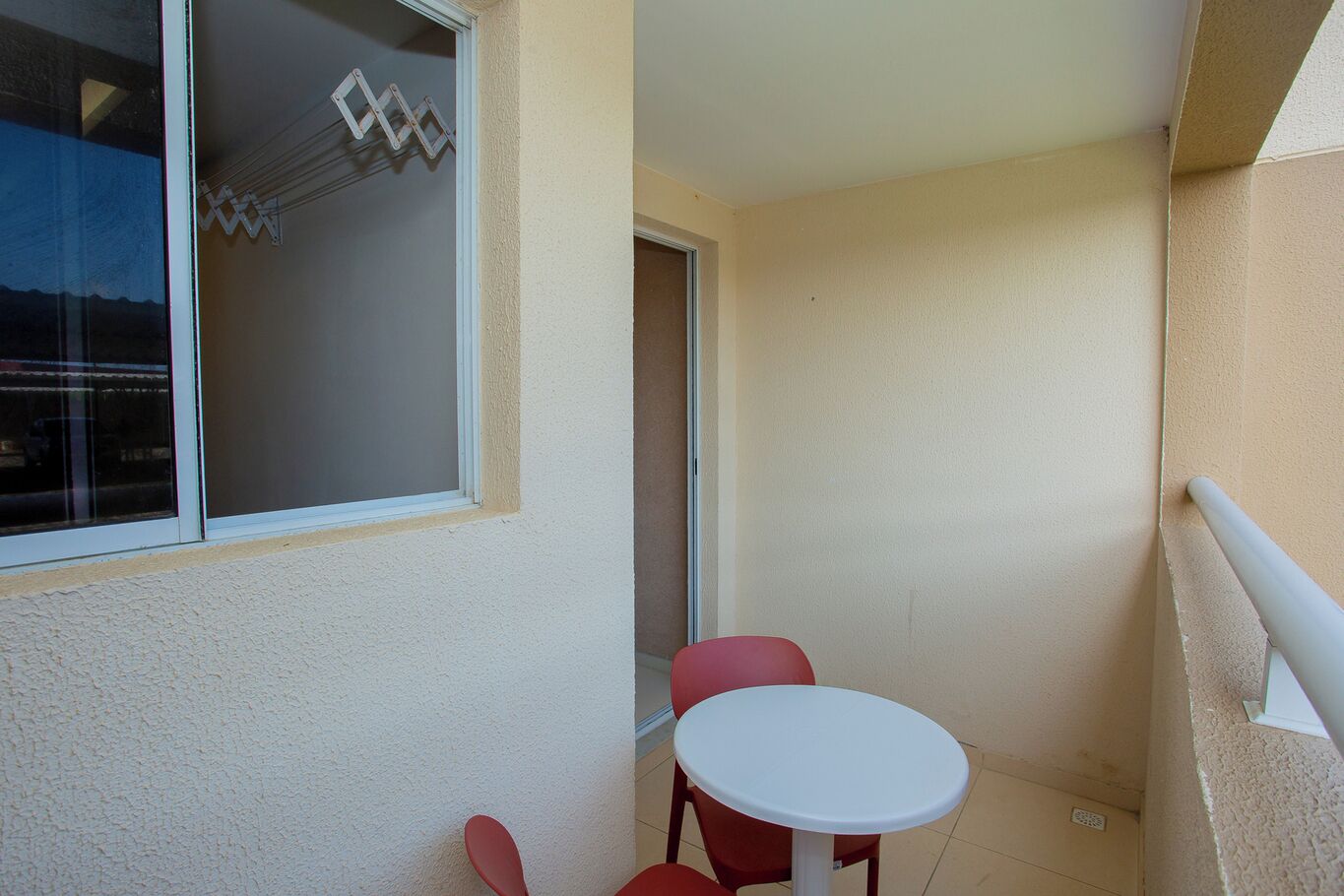 Un petit balcon avec une table blanche et deux chaises rouges, une fenêtre ornée de motifs géométriques et un mur de couleur beige.