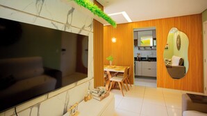 A modern living room with a large flat-screen TV, a dining area with wooden chairs, and a kitchen with wooden cabinets. - Apto com Piscina e Garagem em Barra dos Coqueiros (Barra dos Coqueiros)