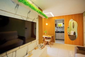 A modern living room with a large flat-screen TV, a dining area with wooden chairs, and a kitchen with wooden cabinets.