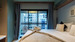 Ett hotellrum med en säng, en stol och utsikt över höga byggnader genom ett stort fönster.