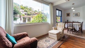 A living room with a large window, a sofa, a chair, a dining table with chairs, and a patterned rug. - Piscina, Lareira e Natureza Incrível - Petrópolis (Petropolis)