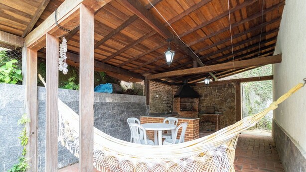 A covered outdoor area with a brick barbecue, white plastic chairs, and a hammock. - Piscina, Lareira e Natureza Incrível - Petrópolis (Petropolis)