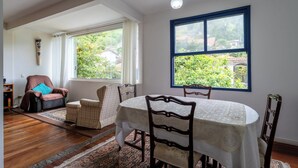 A dining area with a table set for four, surrounded by chairs, a sofa, and a window with a view of greenery. - Piscina, Lareira e Natureza Incrível - Petrópolis (Petropolis)