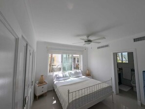 Quarto com cama, ventoinha no teto, janela, mesa de cabeceira e banheiro visível por uma porta aberta.