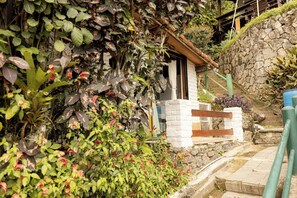 Ein rustikales Steinhaus mit einer Holztreppe, die zu einer weißen Tür führt, umgeben von üppiger Vegetation und einer Steinmauer.