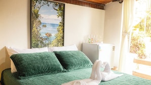 Ein Bett mit grüner Bettwäsche, weißen Kissen und einem Handtuch. Es gibt ein Fenster mit Blick auf viel Grün und eine weiße Wand mit einem gerahmten Bild.