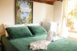 Ein Bett mit grüner Bettwäsche, weißen Kissen und einem Handtuch. Es gibt ein Fenster mit Blick auf viel Grün und eine weiße Wand mit einem gerahmten Bild.