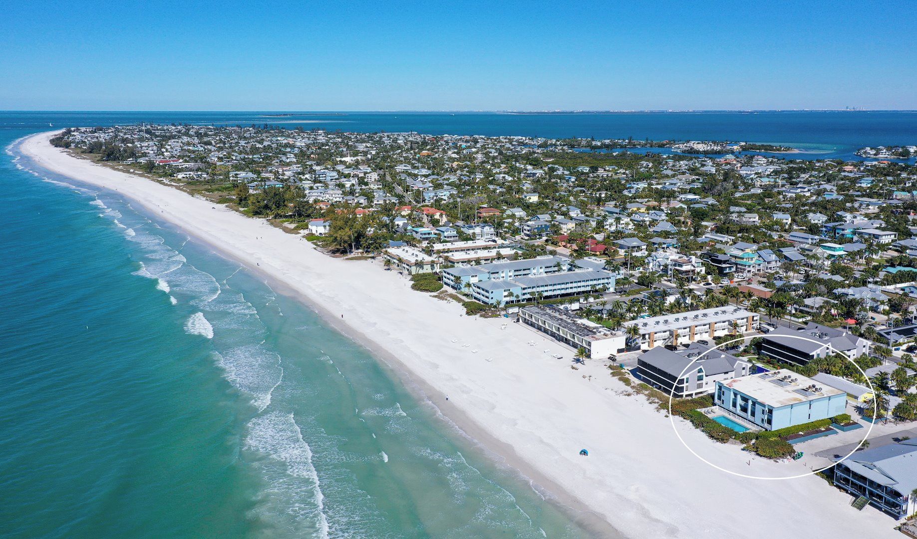 An aerial view of a coastal town with a long sandy beach, residential buildings, and a clear blue sky.