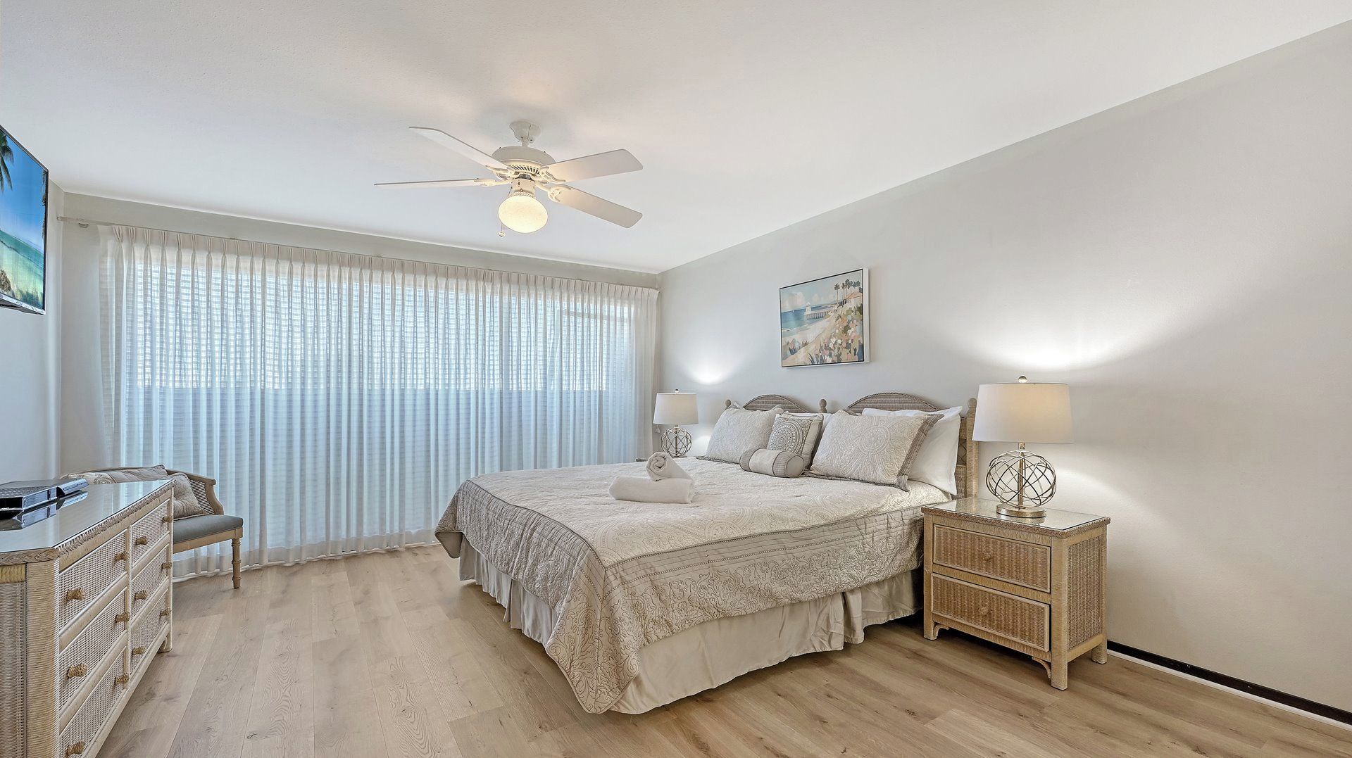 Une chambre à coucher comprenant un lit, des tables de chevet, un ventilateur de plafond et une grande fenêtre ornée de rideaux légers.