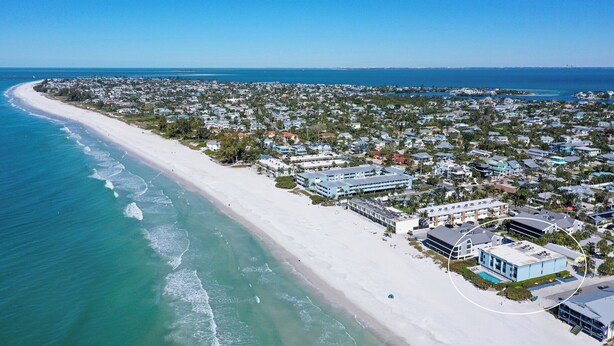 Luftaufnahme einer Küstenstadt mit einem langen Sandstrand, Wohngebäuden und einem klaren, blauen Himmel.