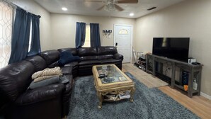 A living room with a leather sofa, a glass coffee table, a television, and a ceiling fan. - Affordable private room King bed in Thonotosassa, shared space (Thonotosassa)