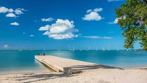 Una spiaggia con un molo in cemento che si protende in acque turchesi calme, circondata da cieli sereni e poche nuvole.