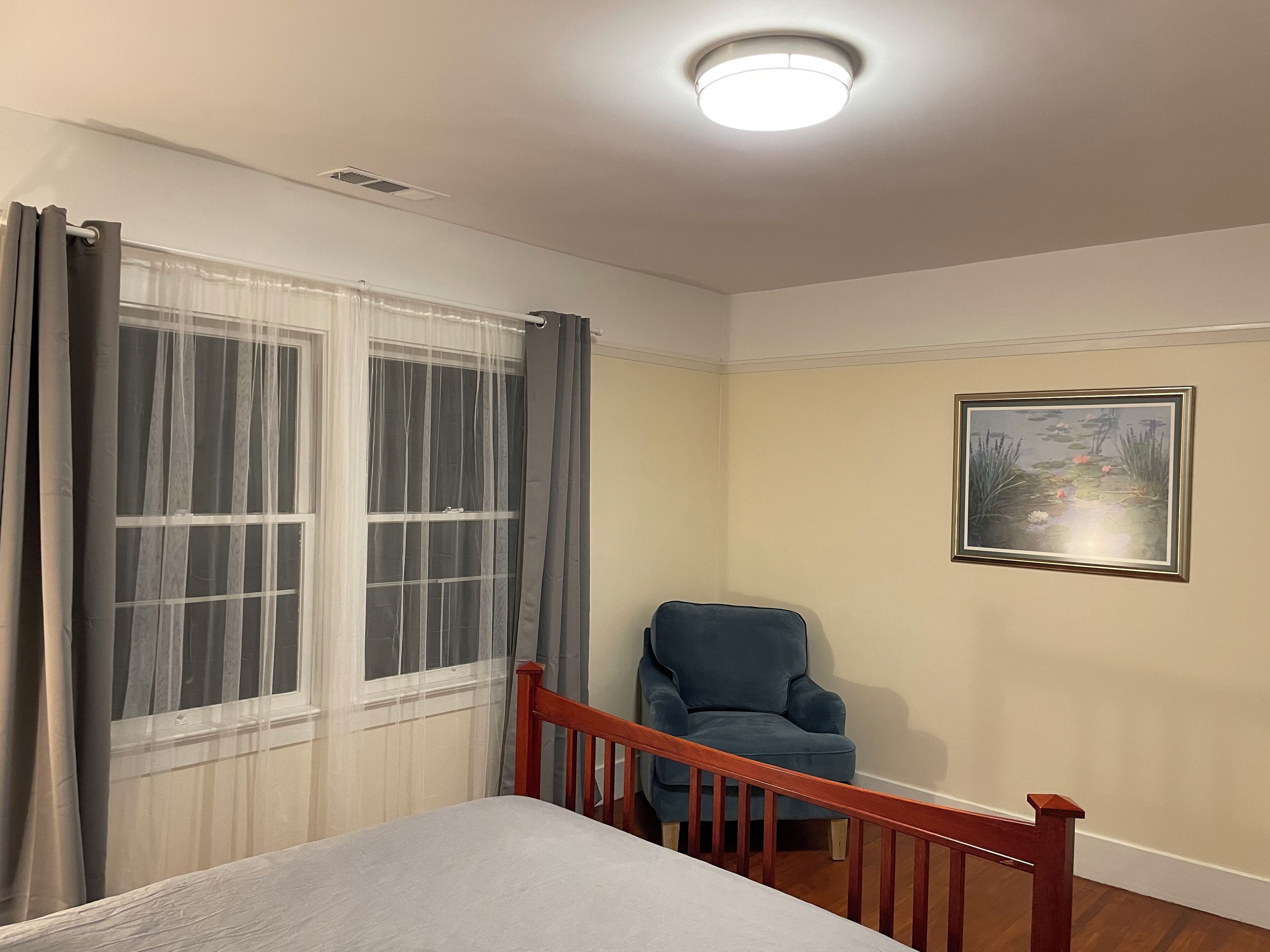 Quarto com cama, cadeira, janela com cortinas, luminária no teto e quadro na parede.