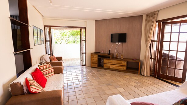 A living room with a sofa, TV, and a sliding glass door leading to an outdoor area. - Casa c/ Piscina a 550m da Praia | Até 10 hóspedes (Lauro de Freitas)