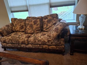 A patterned sofa, a wooden side table with a lamp, and a window with blinds. - Spacious and beautiful 4-bedroom country home with AC in marvelous Bridgeton. (Bridgeton)