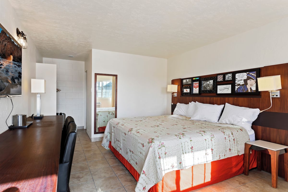 Camera d'albergo con un letto grande, una testiera in legno, un comodino, una scrivania e una sedia. Una finestra permette l'ingresso di luce naturale.