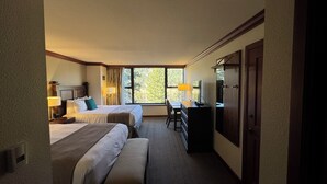 A hotel room with two beds, a desk, a dresser, and a large window with a view of trees.