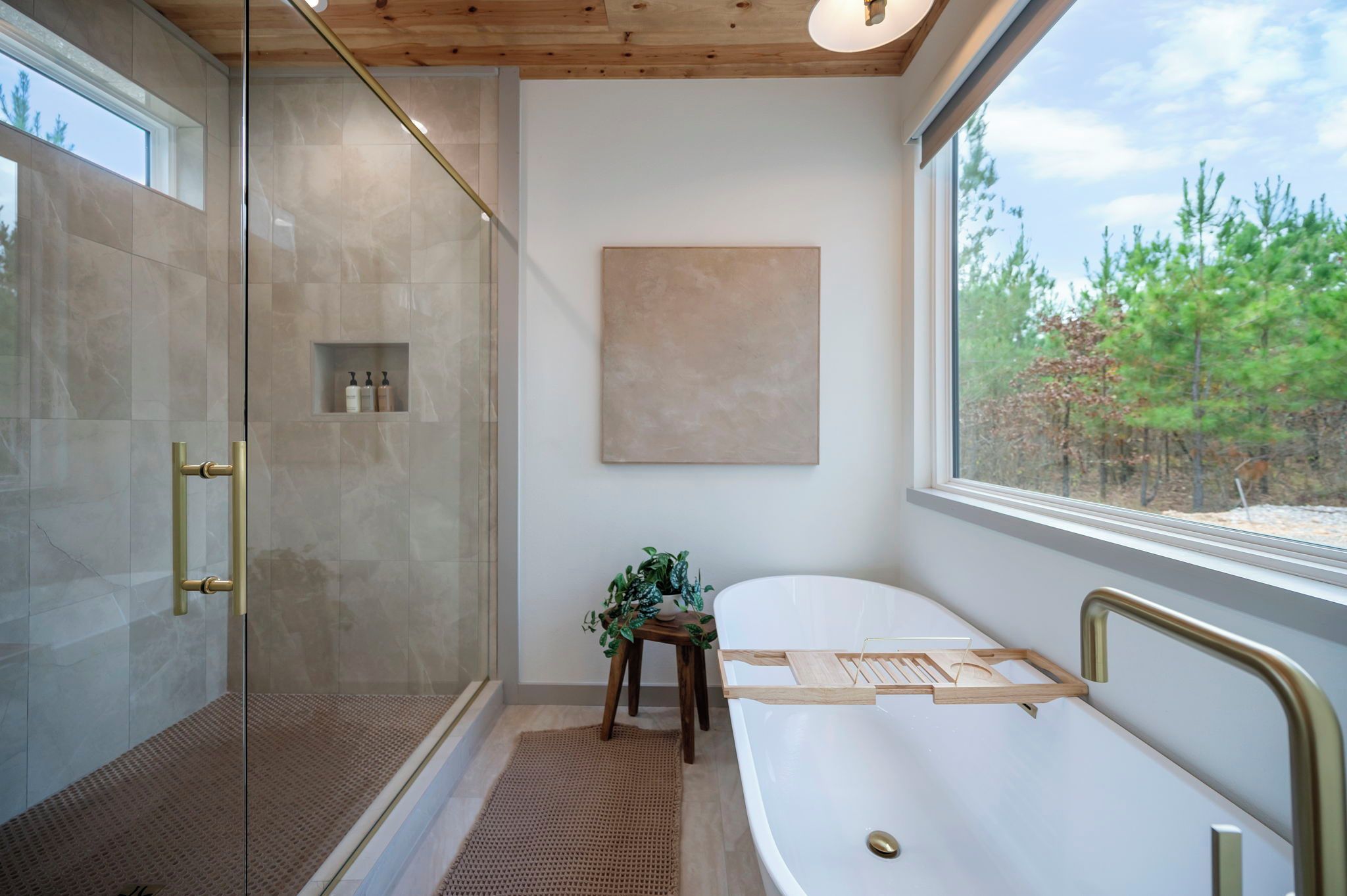 A modern bathroom with a glass shower enclosure, a white bathtub, and a view of trees through a large window.
