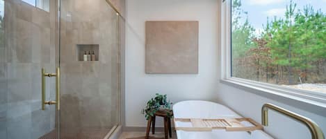 A modern bathroom with a glass shower enclosure, a white bathtub, and a view of trees through a large window.
