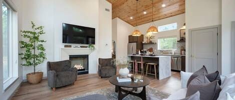 Un salón espacioso con chimenea, asientos cómodos, televisión y una zona de cocina con electrodomésticos modernos e iluminación colgante.