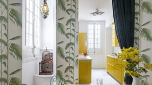 Ein Zimmer mit weiß-gelber Farbgestaltung, ausgestattet mit einem Fenster, einem Vogelkäfig, einer Vase mit gelben Blumen und einer Obstschale auf dem Tisch.