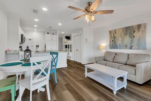 A modern living room with a white sofa, a coffee table, and a dining area with a white table and blue chairs. - Beachside Gem ~ Remodeled Luxury Steps to Sand (Miramar Beach)