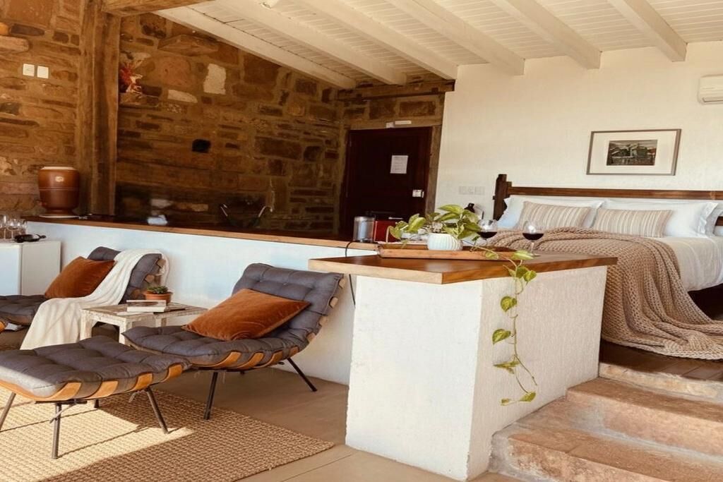 Quarto aconchegante com parede de pedra, cama com cabeceira de madeira, um pequeno espaço de estar com uma poltrona e uma mesa, e janela com vista.