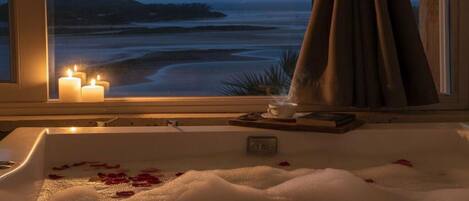 A bathtub with rose petals, candles, and a view of the beach at dusk.