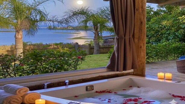 A spa area with a bathtub filled with water and rose petals, a wooden deck, and a view of a lake at night. - HOSPEDARIA PONTA DA PITEIRA (Imbituba)