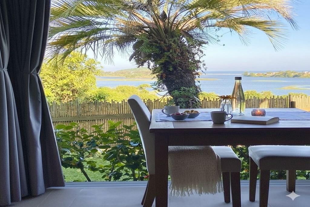 Una mesa de comedor con sillas, una vela y vistas a un paisaje tropical con una palmera y agua.