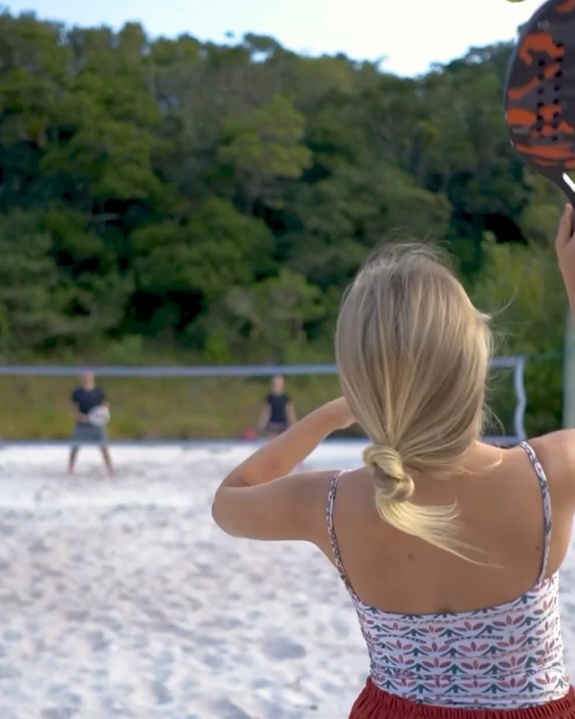 A person in a patterned top and red skirt standing on a sandy beach, holding a volleyball.