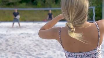 Una persona con una blusa estampada y falda roja parada en una playa arenosa, sosteniendo una pelota de voleibol.