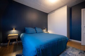 Un dormitori amb un llit, dues taules de nit, una tauleta auxiliar, una porta i una làmpada al sostre.