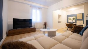 A living room with a large flat-screen TV, a sofa, a coffee table, and a dining area with a chair and a small table. - L'Ecrin Bleu - Spacious - Ideal for couples & professionals - WiFi - Station & Hyper center (Angoulême)
