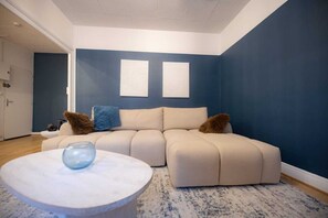 Ein Wohnzimmer mit einem beigefarbenen Ecksofa, zwei an der Wand befestigten Kunstwerken, einem runden Couchtisch mit einem blauen Gegenstand und einer blauen Akzentwand.