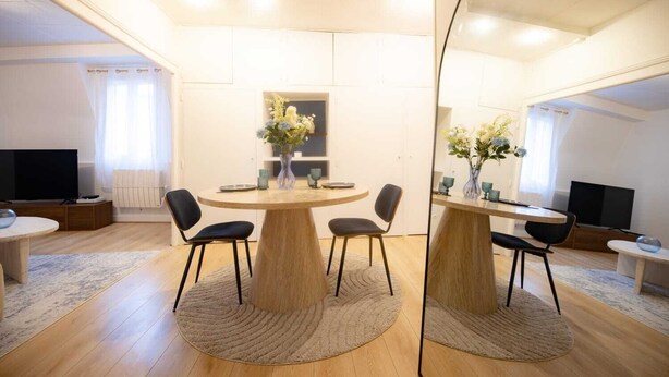 A modern living room with a round wooden table, four black chairs, a flat-screen TV, and a large mirror reflecting the room. - L'Ecrin Bleu - Spacious - Ideal for couples & professionals - WiFi - Station & Hyper center (Angoulême)
