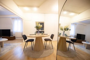 A modern living room with a round wooden table, four black chairs, a flat-screen TV, and a large mirror reflecting the room. - L'Ecrin Bleu - Spacious - Ideal for couples & professionals - WiFi - Station & Hyper center (Angoulême)