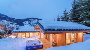 A cozy wooden cabin with a hot tub and a wooden deck, surrounded by snow-covered mountains. - Luxury Chalet with Private Spa & Wellness Area (Megève)