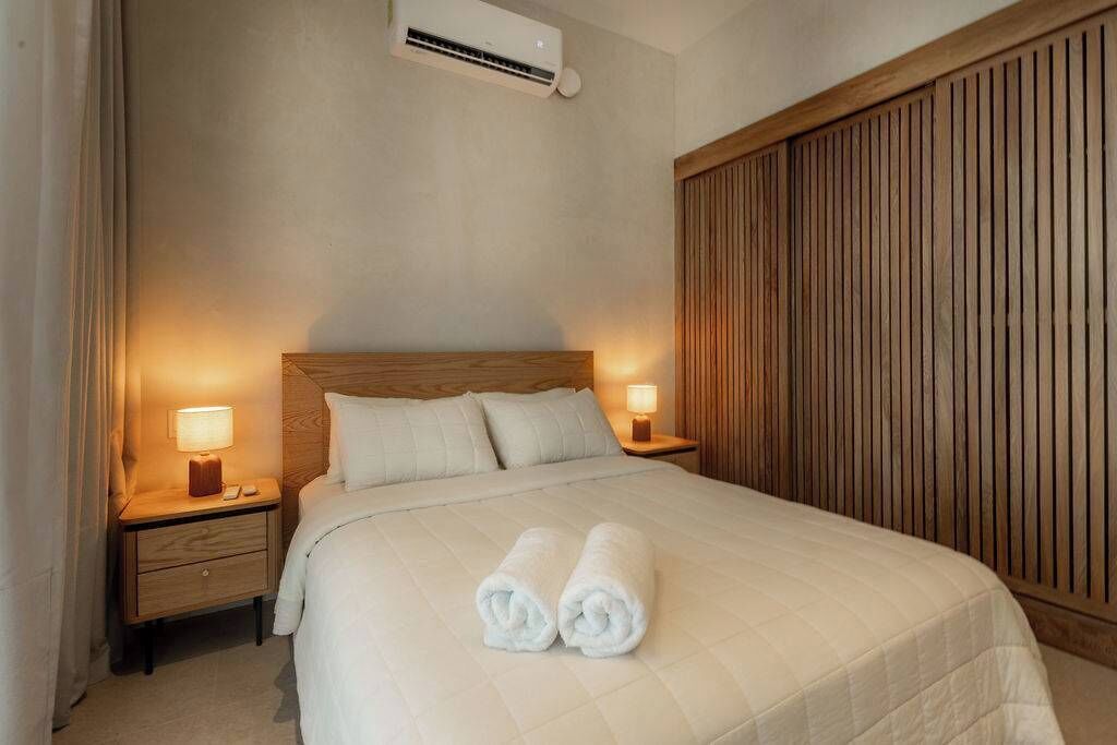 A neatly made bed with white linens and two rolled towels, a wooden headboard, a nightstand with a lamp, and a wooden wardrobe.