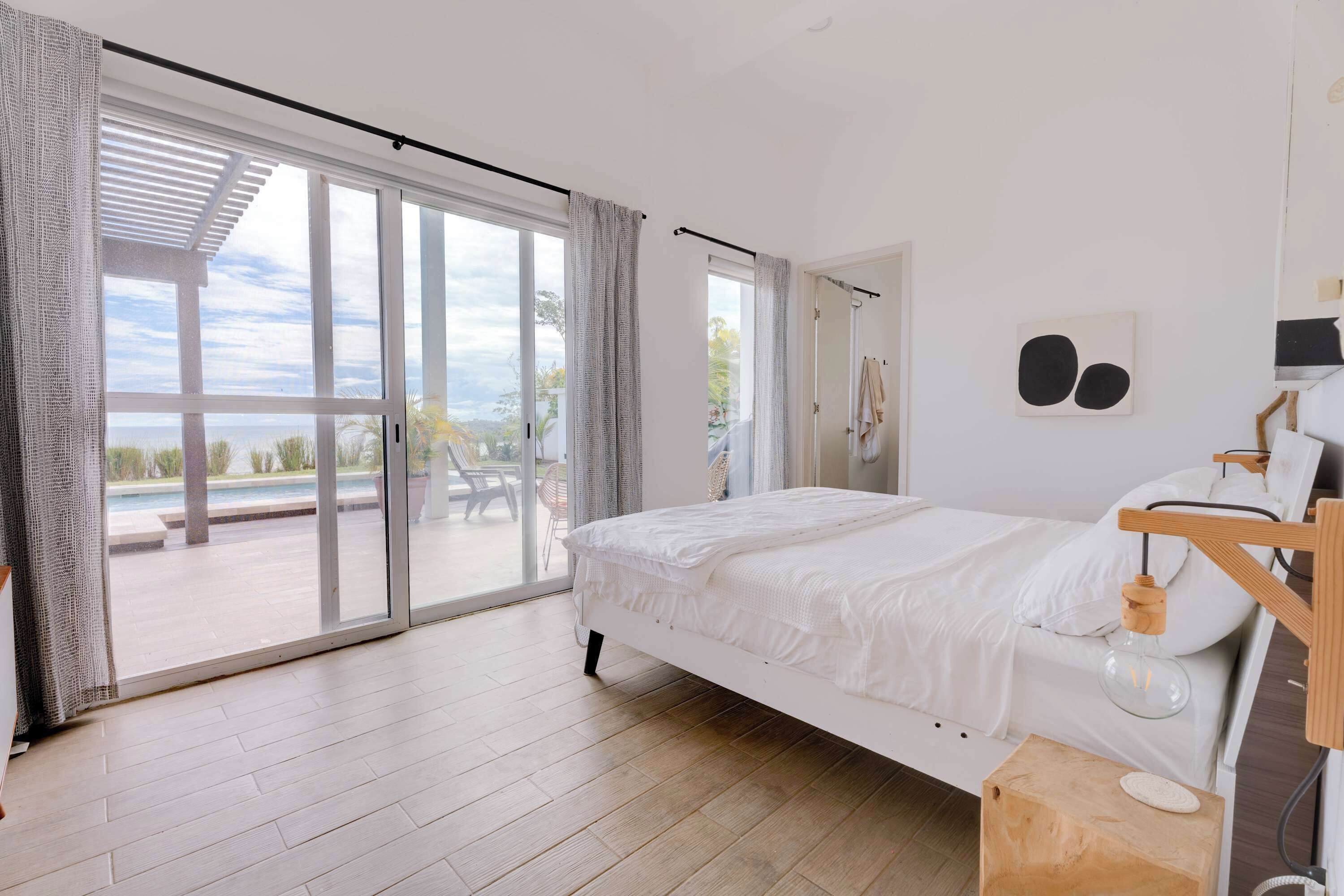 A modern bedroom with a large sliding glass door leading to an outdoor area, a bed with white bedding, and a wooden nightstand.