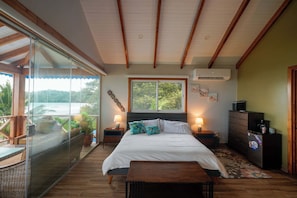 Une chambre avec un grand lit, une porte vitrée donnant sur un balcon, une commode et une petite table avec une lampe.