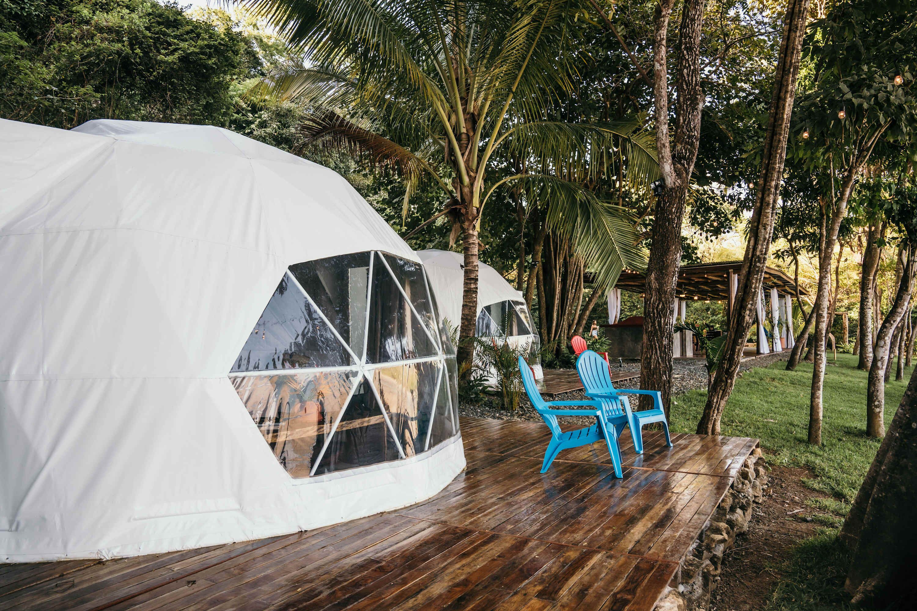 Une tente en forme de dôme dotée d’une fenêtre transparente, d’une terrasse en bois et d’une chaise bleue.