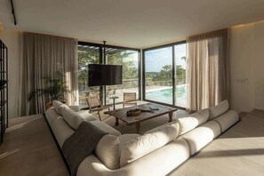 Un salón moderno con un sofá grande, una mesa de centro y un televisor. Se ve una piscina y vegetación al exterior.